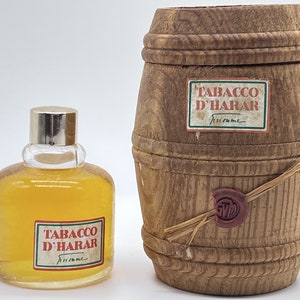 May include: A small bottle of Tabacco D'Harar cologne in a clear glass bottle with a silver cap. The bottle is sitting on a white surface. The cologne is packaged in a wooden barrel-shaped box with a label that reads "Tabacco D'Harar" and a wax seal.