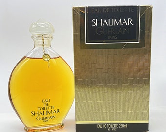 May include: A clear glass bottle of Shalimar Eau de Toilette by Guerlain Paris. The bottle is filled with a golden liquid and has a gold cap. The bottle is sitting on a white surface. The box is gold with a black label that says "Shalimar Guerlain Paris".