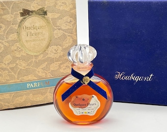 May include: A vintage bottle of Quelques Fleurs Houbigant perfume in a clear glass bottle with a gold cap and a blue ribbon. The bottle is sitting on a blue velvet box with the Houbigant name in gold lettering. The bottle is also in its original box with a lace pattern and the words "Quelques Fleurs Houbigant Parfum" in gold lettering.