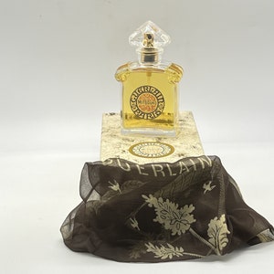 May include: A clear glass bottle of Guerlain Mitsouko perfume with a gold cap and label. The bottle is sitting on a white box with a gold label that says "Guerlain Mitsouko". The box is sitting on a brown fabric with a floral pattern.