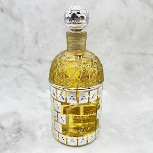 Op de afbeelding: Een parfumfles met een zilver en goud design. De fles heeft een zilveren dop en een goudkleurige body met een textuurpatroon. De onderkant van de fles heeft een zilver en goud design met het woord "EAU" en "GUERLAIN" erop gedrukt.