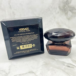 May include: A black box and a perfume bottle. The box is black with white text that reads "VERSACE CRYSTAL NOIR". The perfume bottle is a dark brown square shape with a faceted black cap. The bottle is on a white and gray surface.