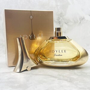 K&ouml;nnte beinhalten: Ein goldfarbener Parf&uuml;mflakon mit dem Aufdruck "IDYLLE" und "Guerlain" auf der Vorderseite. Die Flasche befindet sich neben ihrem Verschluss und einer goldenen Schachtel mit demselben Namen. Der Parf&uuml;mflakon ist mit einer hellgelben Fl&uuml;ssigkeit gef&uuml;llt.