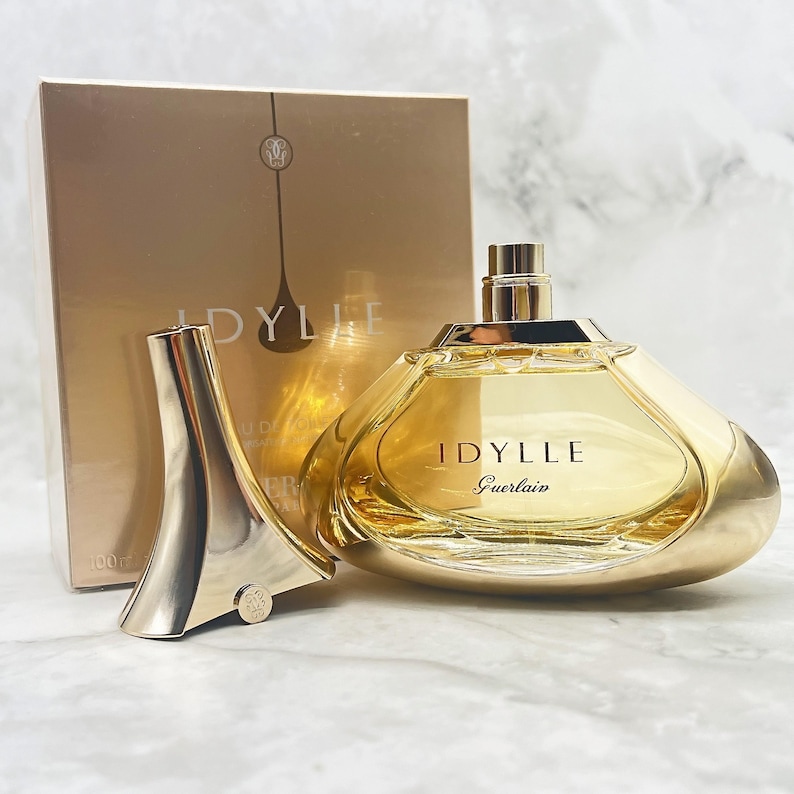 K&ouml;nnte beinhalten: Ein goldfarbener Parf&uuml;mflakon mit dem Aufdruck "IDYLLE" und "Guerlain" auf der Vorderseite. Die Flasche befindet sich neben ihrem Verschluss und einer goldenen Schachtel mit demselben Namen. Der Parf&uuml;mflakon ist mit einer hellgelben Fl&uuml;ssigkeit gef&uuml;llt.