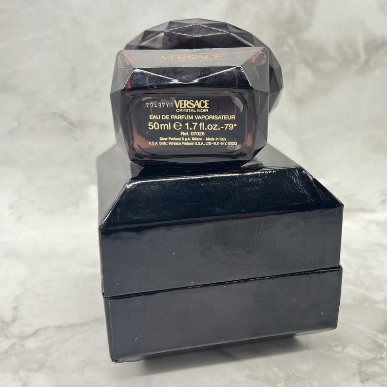 May include: A black Versace Crystal Noir perfume bottle with a faceted cap sits on a black box. The bottle contains 50 ml (1.7 fl. oz.) of perfume. The label includes the text "EAU DE PARFUM VAPORISATEUR".