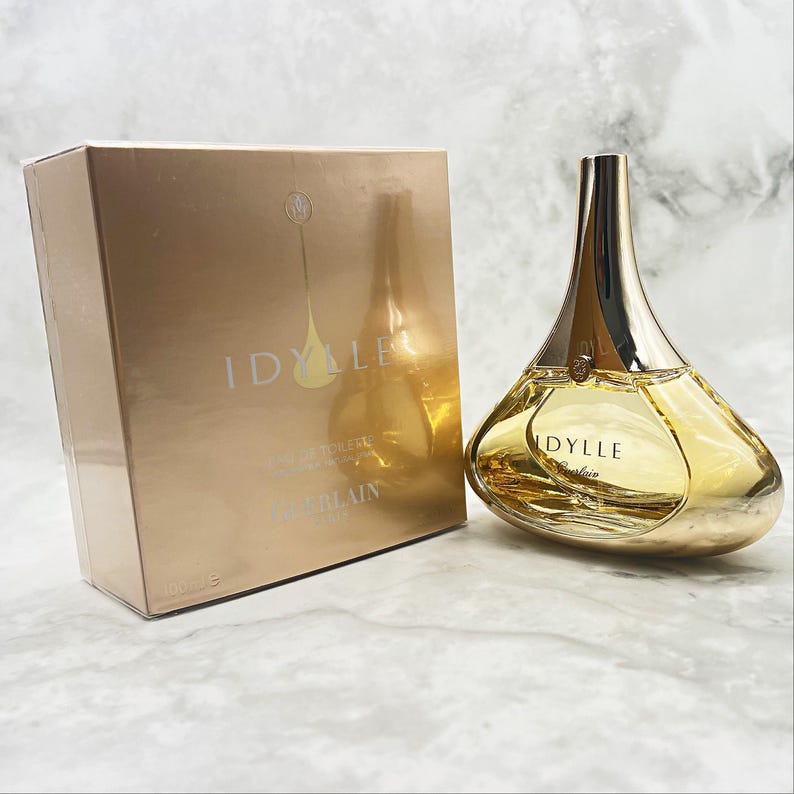 K&ouml;nnte beinhalten: Ein goldfarbenes Parf&uuml;mflaschen- und Box-Set. Die Flasche hat eine einzigartige Tropfenform mit einem goldenen Verschluss und dem Wort "IDYLLE" auf der Vorderseite. Die Box hat eine passende goldene Farbe und tr&auml;gt ebenfalls das Wort "IDYLLE".