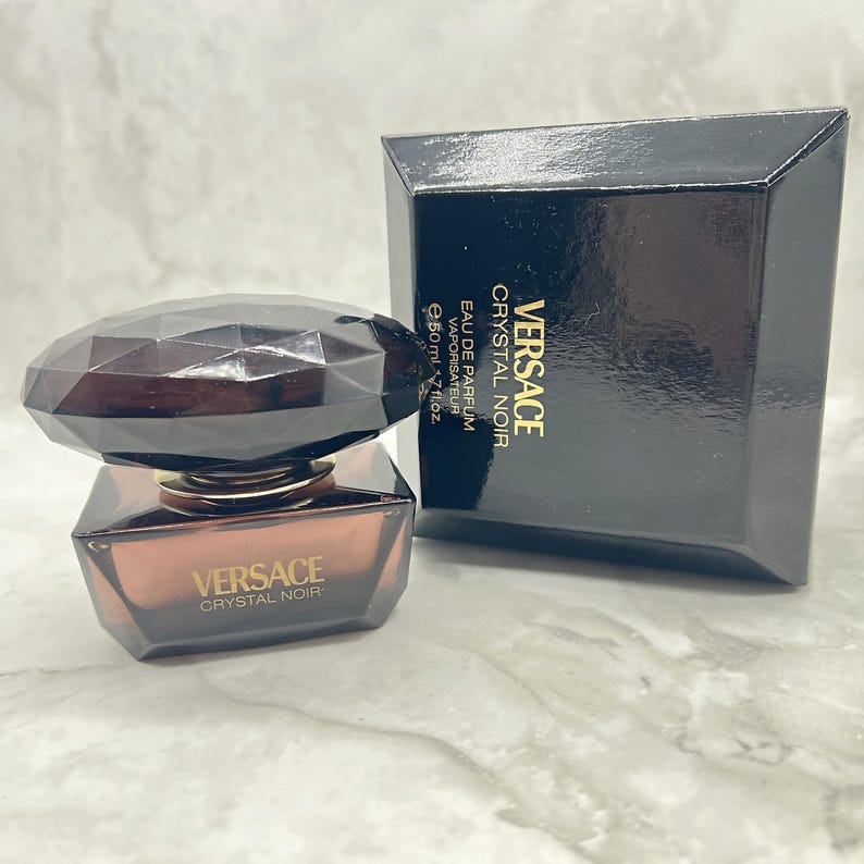 May include: A bottle of Versace Crystal Noir Eau de Parfum with a faceted, dark brown cap and a rectangular, amber-colored glass body. The black box next to it displays the Versace logo and product name.