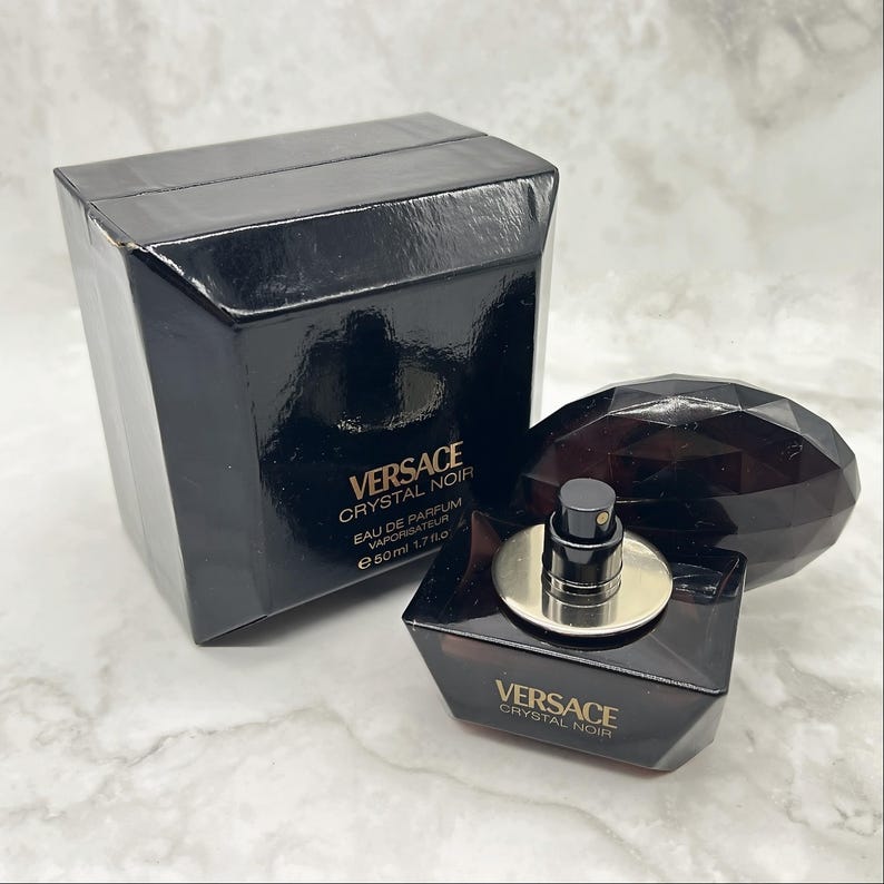 May include: A black Versace Crystal Noir perfume bottle and box set. The perfume bottle is a dark, geometric shape with a silver atomizer. The box is black with the Versace logo and product information. The set includes a 1.7 fl oz (50 ml) bottle.