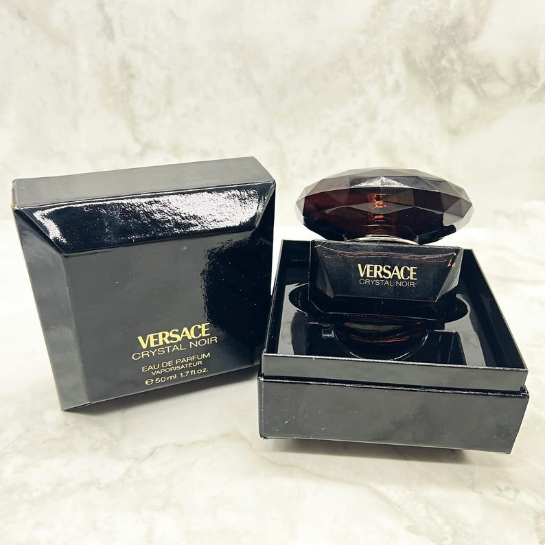 May include: A black perfume bottle and box set. The bottle has a faceted, dark brown cap and a rectangular body. The box and bottle are labeled "Versace Crystal Noir". The box also indicates the perfume is Eau de Parfum, 1.7 fl oz.