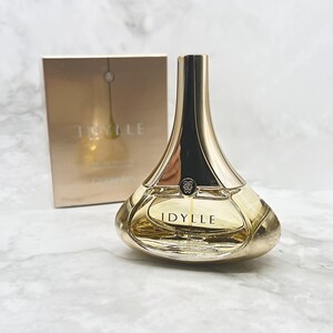May include: A gold-toned perfume bottle with a unique shape, featuring a rounded base and a slender, elongated neck. The bottle is filled with a light amber liquid and is labeled "IDYLLE". A matching box is in the background.