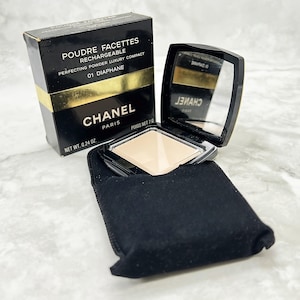 Könnte beinhalten: Ein schwarzes kompaktes Puderetui mit Spiegel und einem schwarzen Samtbeutel. Das Etui trägt den Aufdruck "POUDRE FACETTES RECHARGEABLE PERFECTING POWDER LUXURY COMPACT 01 DIAPHANE CHANEL PARIS POIDS NET 7.6 NET WT. 0.24 OZ."