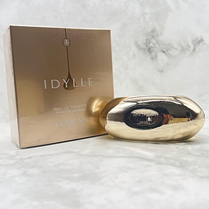K&ouml;nnte beinhalten: Ein goldfarbenes Parf&uuml;mflaschen- und Box-Set. Die rechteckige Box tr&auml;gt den Schriftzug "IDYLLE". Die Flasche ist oval und reflektiert das Licht. Das Set ist f&uuml;r eine 100 ml Flasche.