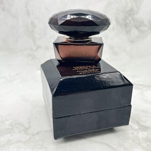 May include: A black Versace Crystal Noir Eau de Parfum bottle with a faceted, dark brown cap. The perfume bottle sits on a black box with the Versace logo and product information. The bottle contains 50 ml / 1.7 fl oz of perfume.