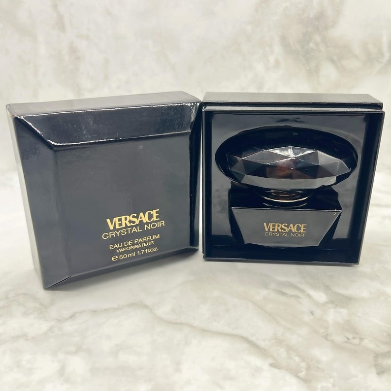 May include: A black Versace Crystal Noir Eau de Parfum bottle and box set. The bottle is a dark, faceted design, and the box features gold lettering. The set includes a 50ml, 1.7 fl oz bottle.