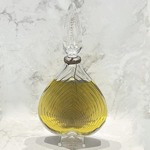 Puede incluir: Una botella de vidrio transparente con un diseño texturizado en forma de corazón. La botella está llena de un líquido amarillo y tiene un tapón de vidrio transparente con la palabra "CHAMADE" grabada.