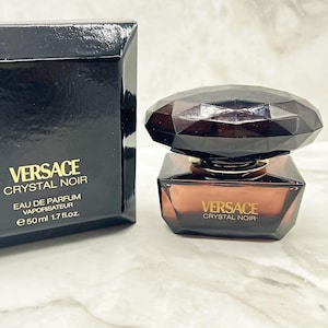 Könnte beinhalten: Eine schwarze Parfümbox und Flasche von Versace Crystal Noir. Die Flasche hat eine dunkelbernsteinfarbene Farbe mit einem facettierten, dunkelbraunen Deckel. Auf der Box und der Flasche sind das Versace-Logo und der Produktname abgebildet. Die Flasche enthält 50 ml.