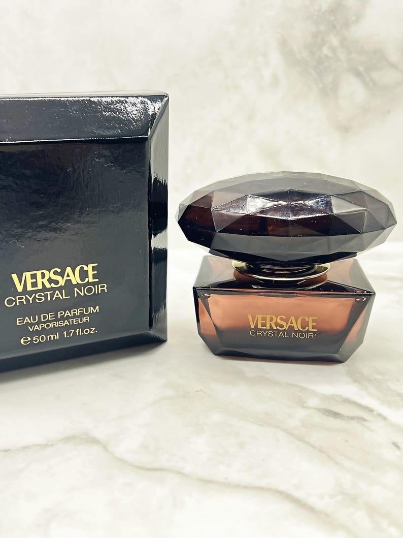 May include: A black perfume box and bottle of Versace Crystal Noir. The bottle is a dark amber color with a faceted, dark brown cap. The box and bottle both feature the Versace logo and product name. The bottle contains 50 ml or 1.7 fl. oz.