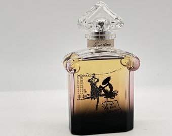 May include: A clear glass bottle of Guerlain perfume with a black and gold label. The label features a black and white illustration of a woman in a dress and a hat. The text on the label reads "La Petite Robe Noire".