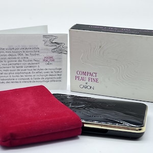 May include: A black compact powder case with a gold trim and a red velvet pouch. The case has a swirling pattern and the text "COMPACT PEAU FINE CARON" is printed on the front. The text "POUDRE PEAU FINE CARON" is printed on the back of the case.