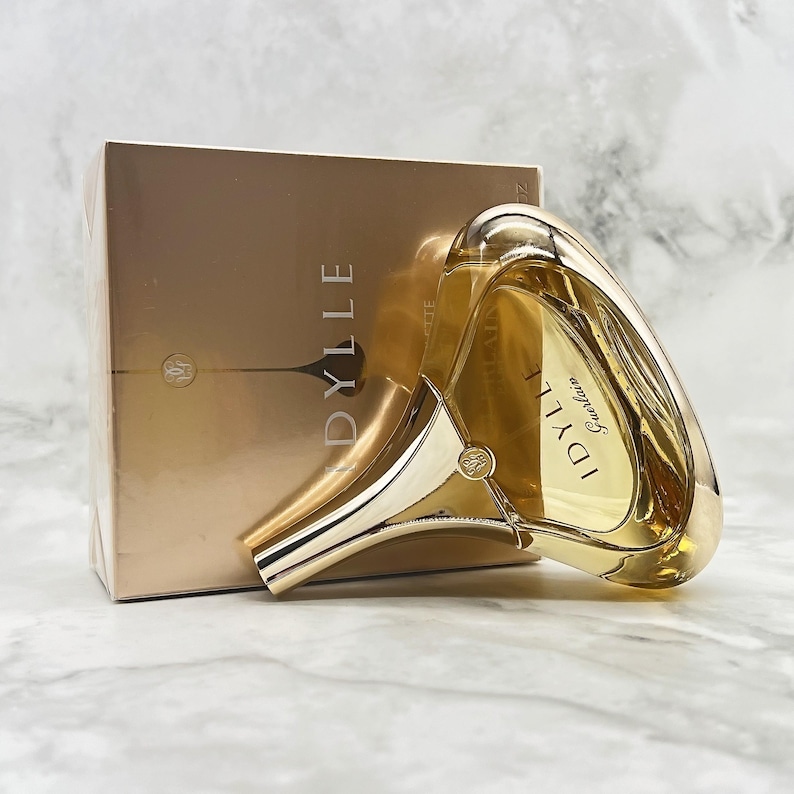 K&ouml;nnte beinhalten: Ein goldfarbenes Parf&uuml;mflaschen- und Box-Set. Die Flasche hat ein einzigartiges, geschwungenes Design mit dem Aufdruck "IDYLLE" und "Guerlain". Die Box ist in passender Goldfarbe gehalten und tr&auml;gt den Aufdruck "IDYLLE".