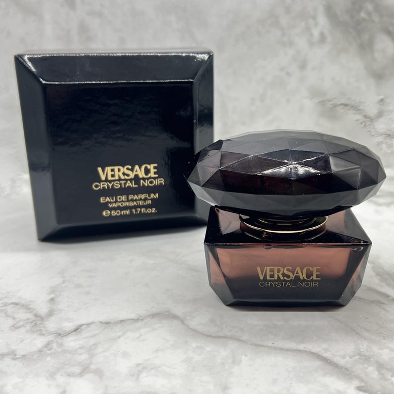 May include: A black Versace Crystal Noir perfume bottle and box. The bottle is a dark brown, geometric shape with a faceted cap. The box is black with gold lettering. The bottle contains 50ml or 1.7 fl oz of perfume.