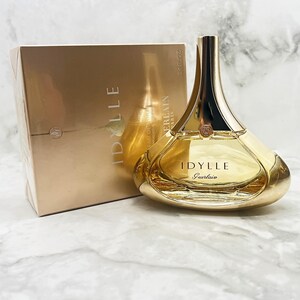 K&ouml;nnte beinhalten: Ein goldfarbenes Parf&uuml;mflaschen- und Box-Set. Die Flasche hat eine einzigartige, geschwungene Form mit einem goldenen Verschluss und dem Namen "IDYLLE Guerlain" auf der Vorderseite. Die Box ist in passendem Gold gehalten.
