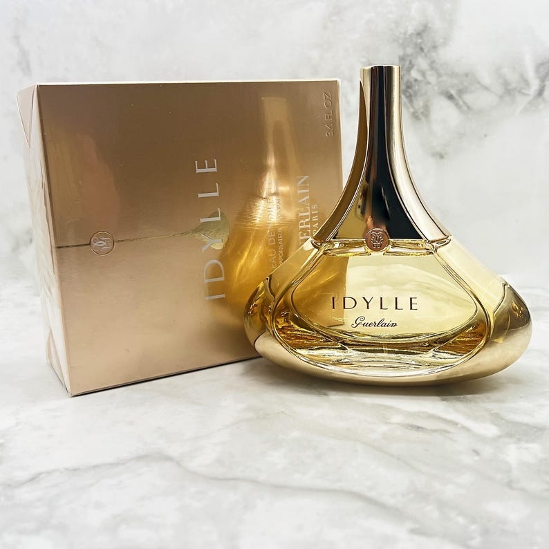 K&ouml;nnte beinhalten: Ein goldfarbenes Parf&uuml;mflaschen- und Box-Set. Die Flasche hat eine einzigartige, geschwungene Form mit einem goldenen Verschluss und dem Namen "IDYLLE Guerlain" auf der Vorderseite. Die Box ist in passendem Gold gehalten.