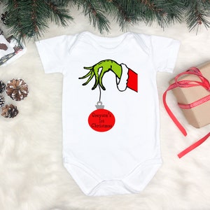 May include: White baby onesie with a green Grinch hand holding a red Christmas ornament that says "Greyson's 1st Christmas".