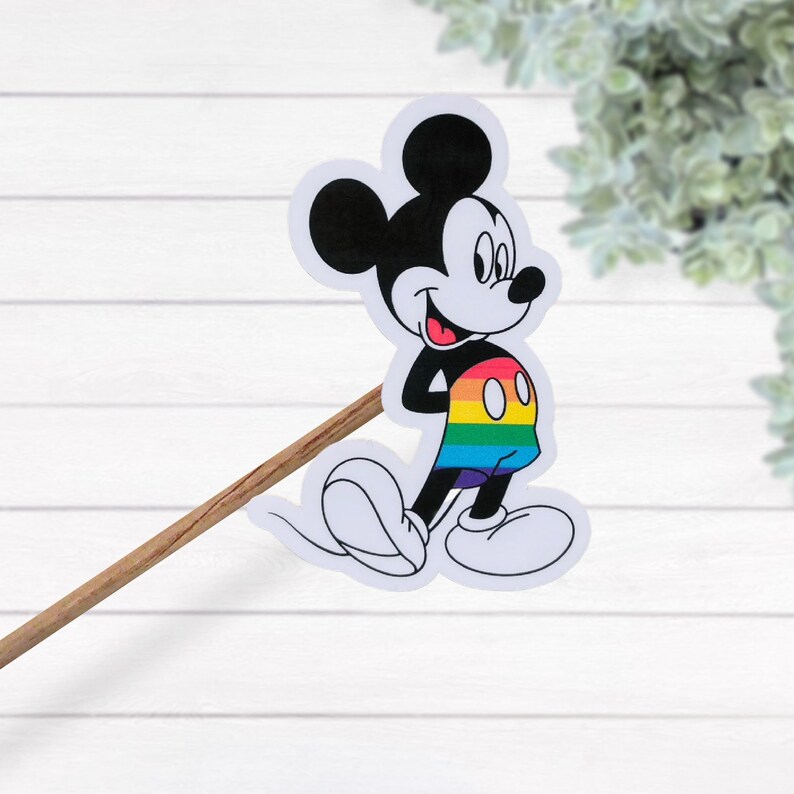 May include: A sticker of Mickey Mouse wearing rainbow shorts. The sticker is on a wooden stick.
