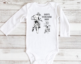 May include: White long-sleeve baby bodysuit with a black graphic of two people riding dirt bikes. The text "Daddy's Future Riding Buddy" is printed on the bodysuit.