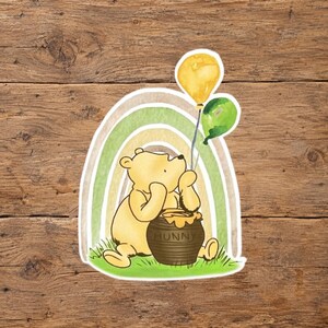 Può includere: Un adesivo raffigurante un orso giallo seduto sull'erba verde davanti a un arcobaleno dai toni tenui. L'orso tiene in mano un palloncino giallo e uno verde, appoggiandosi a un vasetto di miele marrone con la scritta HUNNY sul davanti.