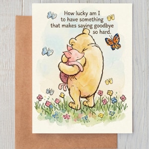 Op de afbeelding: Een wenskaart met een aquarelillustratie van een gele beer die een roze biggetje knuffelt in een veld met kleurrijke bloemen en vlinders. De tekst luidt, How lucky am I to have something that makes saying goodbye so hard.