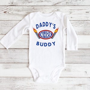 May include: White long-sleeve baby bodysuit with the text "Daddy's NHRA Buddy" in blue and red lettering with a graphic of a red oval with the text "NHRA" in blue and two flames on either side of the oval.