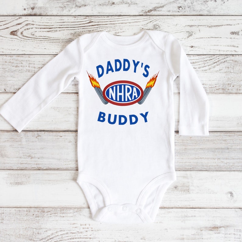May include: White long-sleeve baby bodysuit with the text "Daddy's NHRA Buddy" in blue and red lettering with a graphic of a red oval with the text "NHRA" in blue and two flames on either side of the oval.