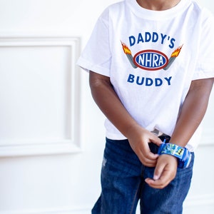 May include: A white t-shirt with the text "Daddy's NHRA Buddy" and a graphic of a racing car with flames. The shirt is being worn by a child.