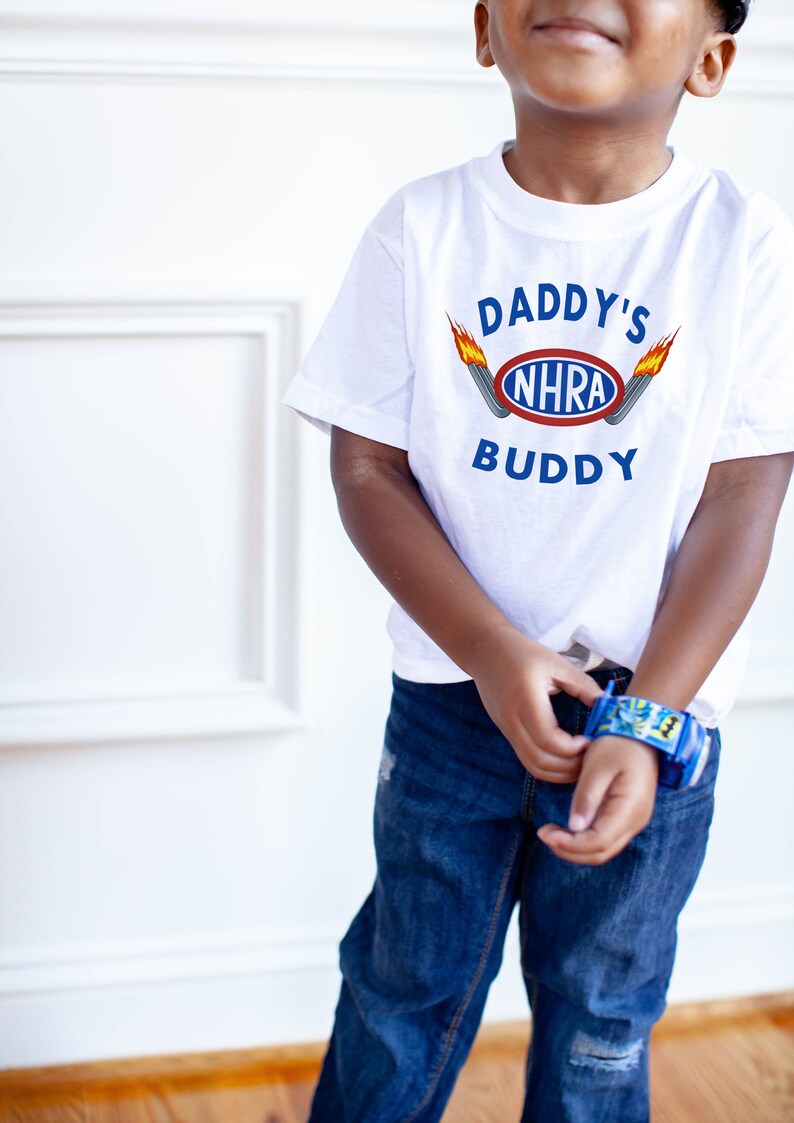 May include: A white t-shirt with the text "Daddy's NHRA Buddy" and a graphic of a racing car with flames. The shirt is being worn by a child.