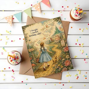May include: A rectangular greeting card featuring an illustration of a person in a blue gingham dress walking with a small dog on a yellow brick road. The text reads, I thought I smelled birthday cake! with signs pointing toward a party.