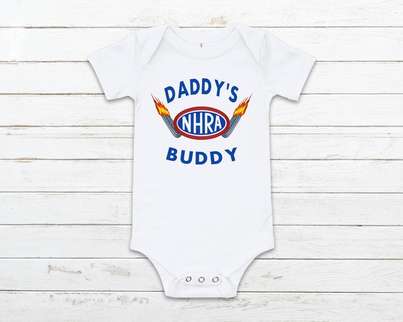 May include: White baby onesie with the text "Daddy's NHRA Buddy" in blue and red lettering with a red oval with the text "NHRA" in blue. The onesie has two flames on either side of the oval.
