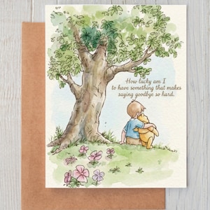 Puede incluir: Una tarjeta de felicitación con una ilustración en acuarela de un niño abrazando a un oso amarillo bajo un gran árbol. El texto dice, How lucky am I to have something that makes saying goodbye so hard. La tarjeta está sobre un sobre marrón.