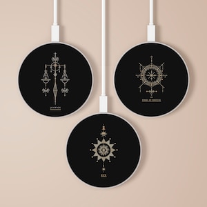 Puede incluir: Tres cargadores inalámbricos negros con diseños geométricos dorados y texto. Los diseños son Justicia, Rueda de la Fortuna y Sol.