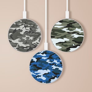 Puede incluir: Tres cargadores inalámbricos redondos con diferentes patrones de camuflaje. Los cargadores son blancos con un contorno negro y están colgados de cables blancos sobre un fondo marrón claro.