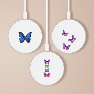 Puede incluir: Tres bases de carga inalámbricas blancas con diferentes diseños de mariposas. La primera base tiene una sola mariposa azul, la segunda base tiene tres mariposas moradas y la tercera base tiene cinco mariposas en una línea vertical, incluyendo mariposas moradas, azules, amarillas y rosas.