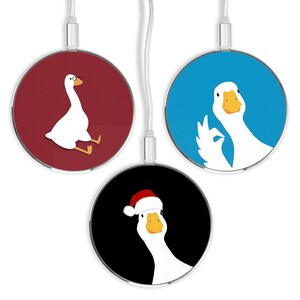 Puede incluir: Tres bases de carga inalámbrica con diferentes ilustraciones de un ganso blanco. La primera base tiene un fondo rojo y el ganso lleva gafas y está sentado. La segunda base tiene un fondo azul y el ganso está haciendo un pulgar hacia arriba. La tercera base tiene un fondo negro y el ganso lleva un gorro de Papá Noel.