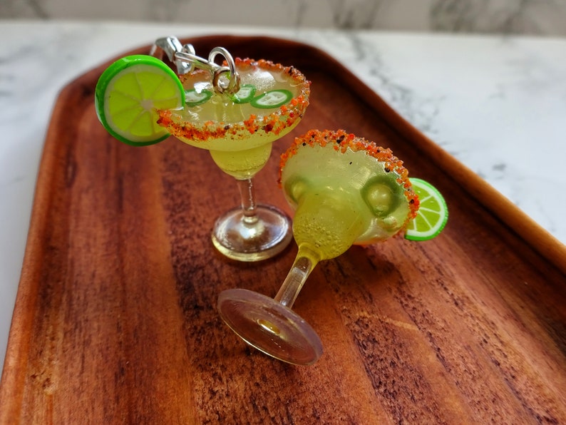 Earrings shaped like a margarita cocktail, a take on a spicy margarita. The cup contains spicy red and orange coloured sand on the rim of the glass. Faux lime fruit slice wedged on the edge with faux jalapenos inside a white/yellow margarita liquid.