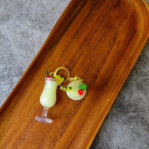 May include: A pair of gold-tone earrings with a tropical drink charm. One earring features a glass of pineapple juice with a cherry and a pineapple wedge. The other earring features a coconut with a cherry and a green leaf.
