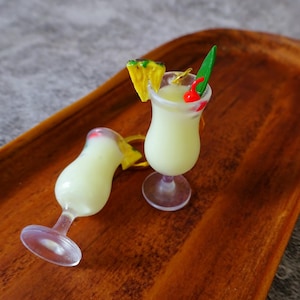 May include: Two miniature plastic cocktail glasses filled with a white liquid, one with a pineapple slice and a green leaf garnish, the other with a red cherry garnish. The glasses are on a wooden tray.