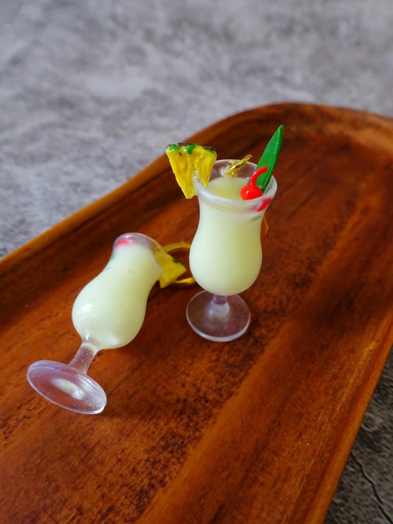 May include: Two miniature plastic cocktail glasses filled with a white liquid, one with a pineapple slice and a green leaf garnish, the other with a red cherry garnish. The glasses are on a wooden tray.