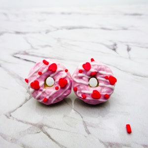 Puede incluir: Dos donuts glaseados de color rosa y blanco con chispas rojas en forma de corazón. Los donuts están sobre una superficie de mármol blanco.