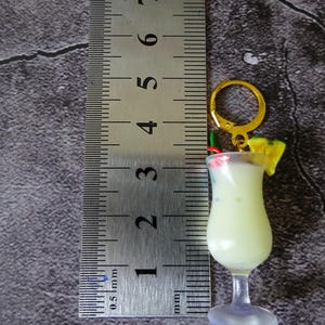 May include: A yellow and white plastic keychain shaped like a cocktail glass with a pineapple, red and green straws, and a gold ring. The keychain is about 2 inches tall and is measured against a ruler.