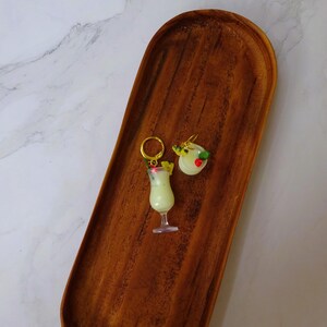 May include: A pair of gold earrings with a white and green cocktail drink charm and a white and red pineapple charm.